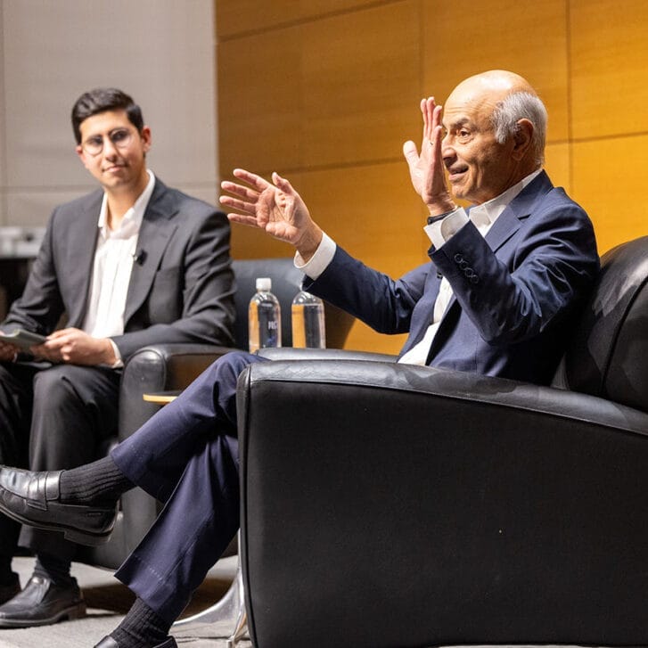 An alumnus and a student sit in cushioned chairs on a stage. Both of them are in suits.