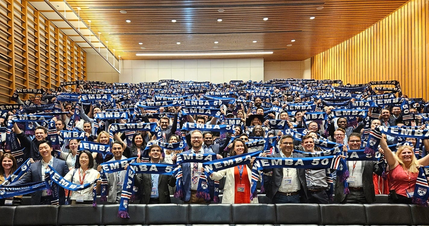 Hundreds of Wharton students stand in auditorium rows holding up blue scarves with the words 
